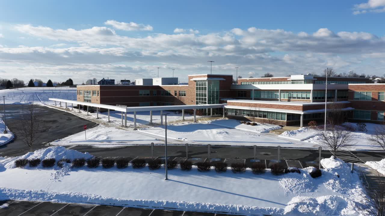 Aerial view of snow-covered school campus in Manheim, Pennsylvania, showing modern buildings, empty parking areas and winter landscape under blue skies. Quiet suburban American winter scene