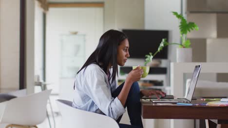 Mixed-race-woman-using-laptop-writing-in-notebook-drinking-coffee-working-from-home