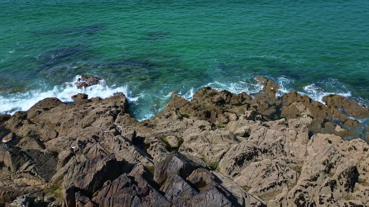 Drone reveals jagged rocks and waves crashing before reaching green vegetation and coastal cave - France