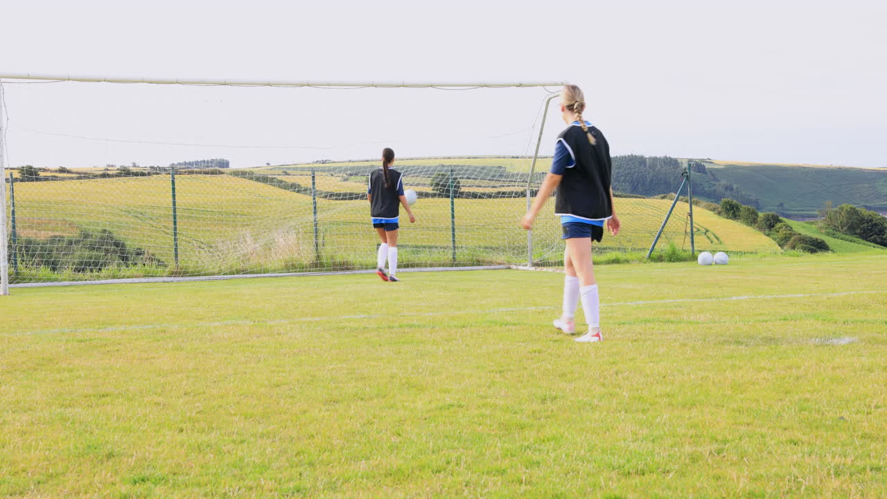 Female soccer players wearing black vests, practicing shooting at goal and passing ball on pitch