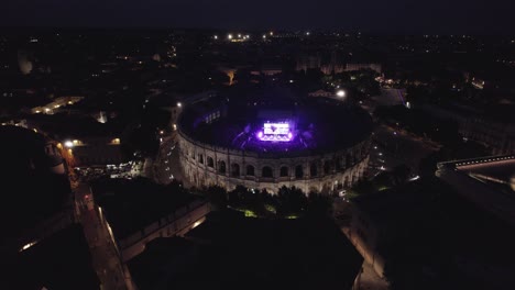 Drone-Para-Prestar-Lento-En-Las-Arenas-De-Nîmes-En-Medio-De-La-Noche,-La-Gente-Está-Viendo-El-Concierto-Y-Hay-Luces-De-Varios-Colores