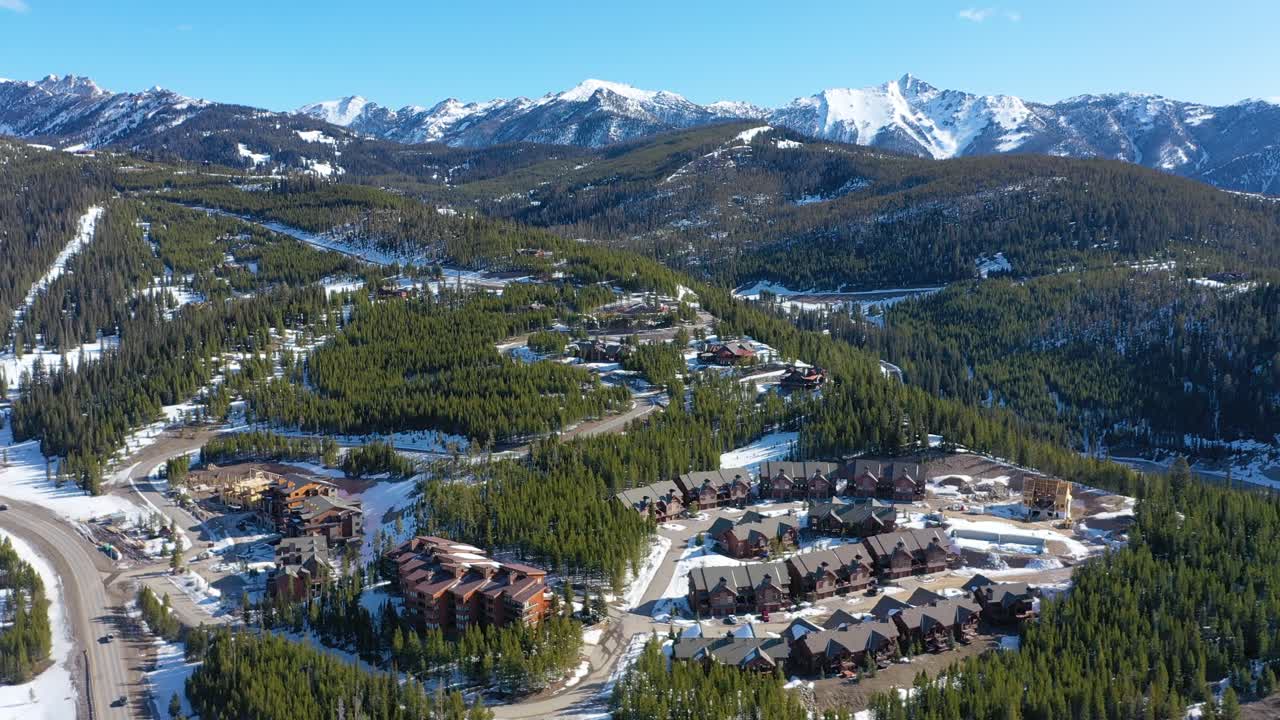 Drone glides above Big Sky, Montana in winter, revealing cozy condos, snow-lined roads, and pine forests throughout the mountain town in a peaceful high-elevation alpine setting