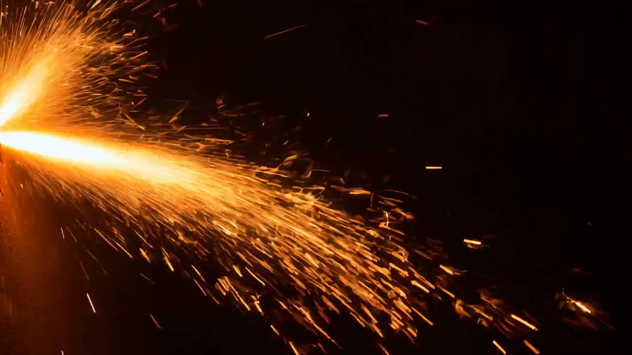 Close-up view of glowing orange sparks flying diagonally across a black background, creating a dramatic high-contrast scene with motion trails, scattered light