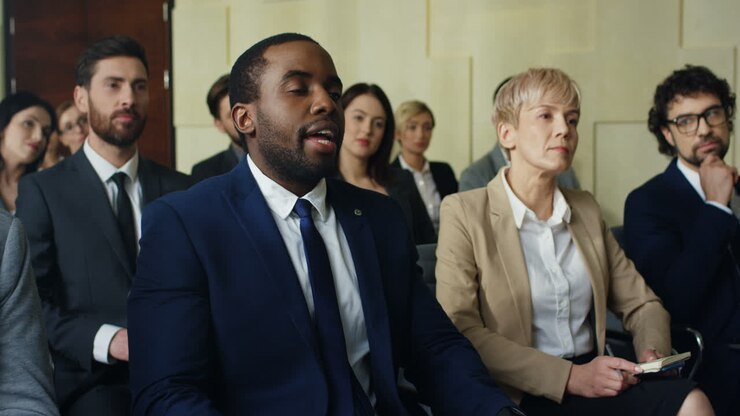 African American young businessman sitting on a chair among people in a conference room and rising his hand to ask a question
