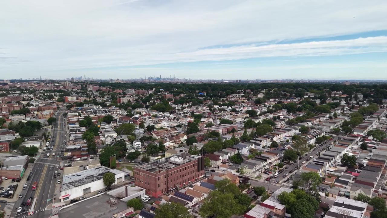 Tomada aérea de alto ângulo sobrevoando um bairro residencial no Queens, Nova York, mostrando moradias densas, ruas com tráfego e o icônico horizonte de Manhattan visível no horizonte
