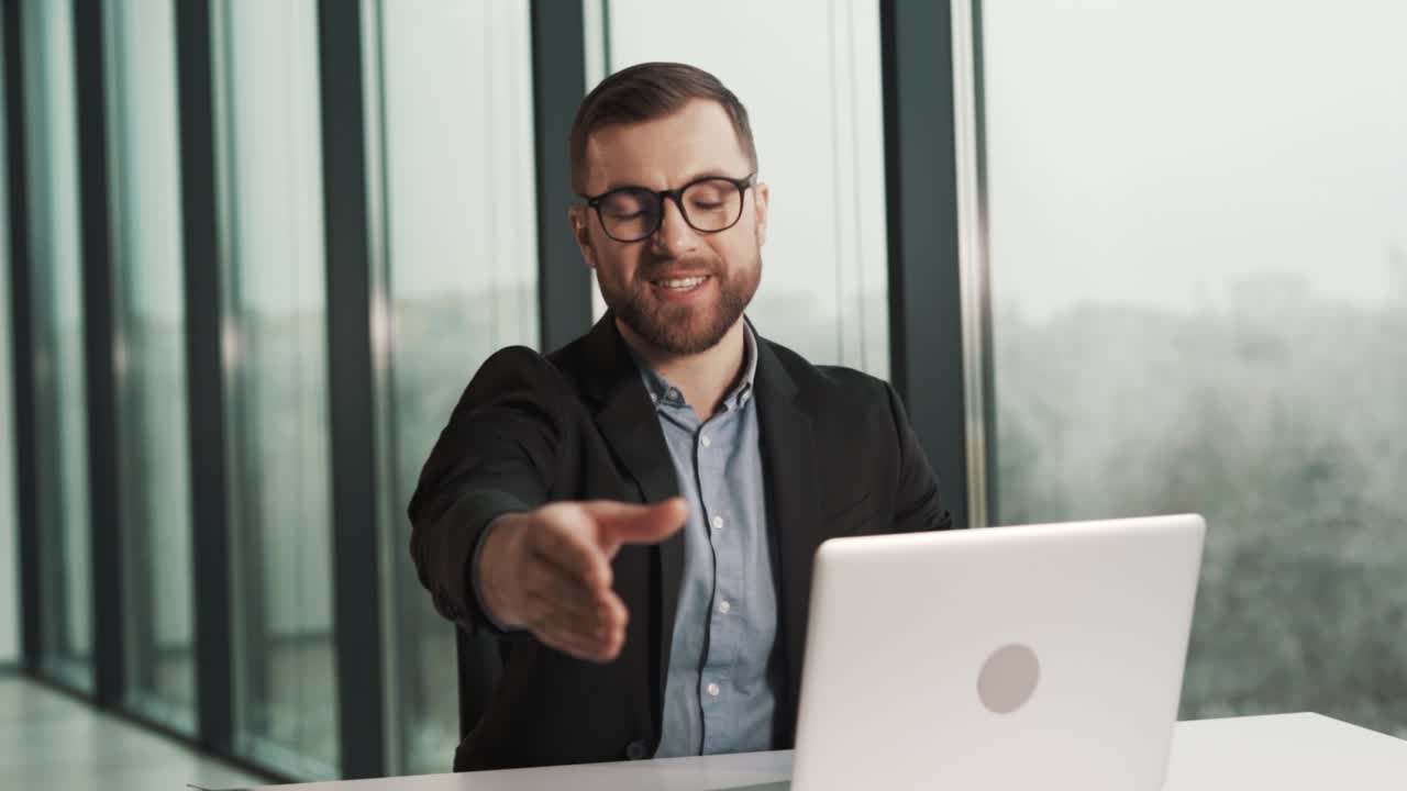 un hombre sonriente con chaqueta y gafas le da la mano a un empleado obvio de la empresa en la oficina sentado en una mesa con una laptop