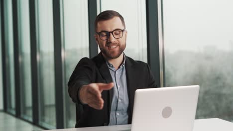 a smiling man in a jacket and glasses shakes hands with an obvious employee of the company in the office sitting at a table with a laptop