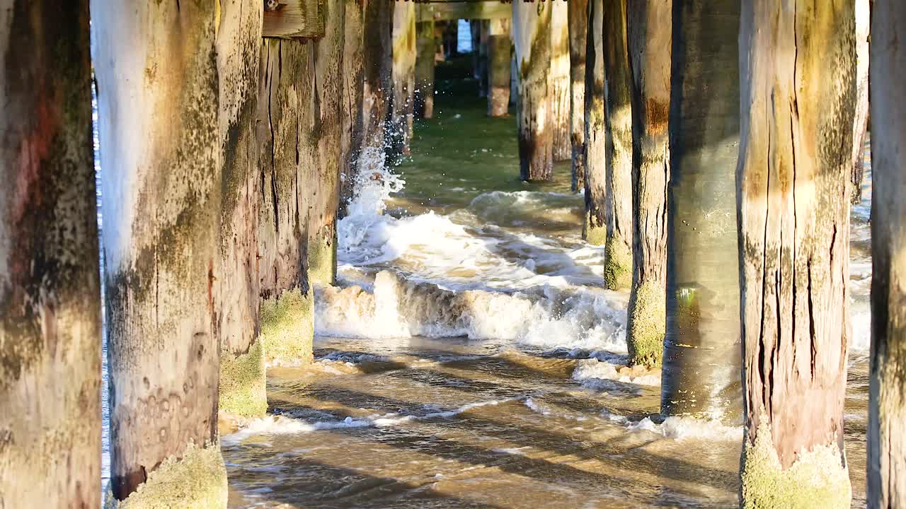 Dynamic waves crash under a weathered wooden bridge in Bellarine, Victoria. Sunlight highlights the textured pillars and moving water
