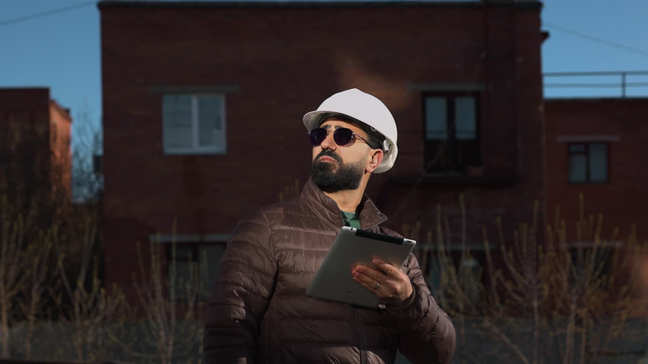 Mand med hat inspicerer tag med tablet og solbriller, dunjakke mod industrielle murstensbygninger