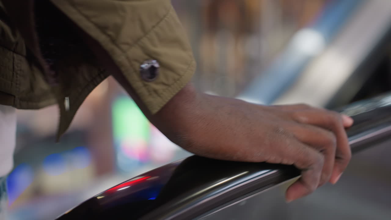 Close up of human hand touching escalator rail inside modern indoor space with glossy surface and blurred colorful reflections creating vibrant city mood while escalator steps move in soft background