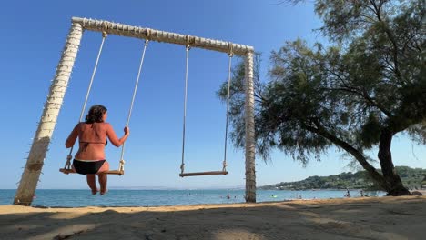 Tiefwinkel-Rückansicht-Einer-Frau-Im-Sommerurlaub,-Die-Sich-Entspannt-Und-Auf-Einer-Seilschaukel-Schwingt,-Während-Sie-Auf-Ruhige-See-Blickt