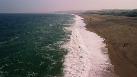 A-drone-shot-tracking-along-a-beach-in-Devon-Uk-people-are-walking-on-the-beach-waves-are-on-the-left-sand-is-on-the-right