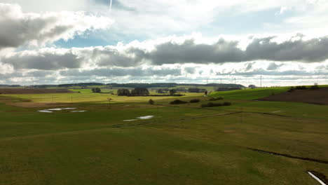 Grassy-field-with-water-patches,-rolling-hills-with-trees,-expansive-rural-backdrop,-dynamic-cloudy-sky