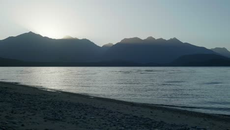 Sunlight-pierces-through-mountains,-casting-ethereal-rays-over-a-tranquil-lake-at-dawn