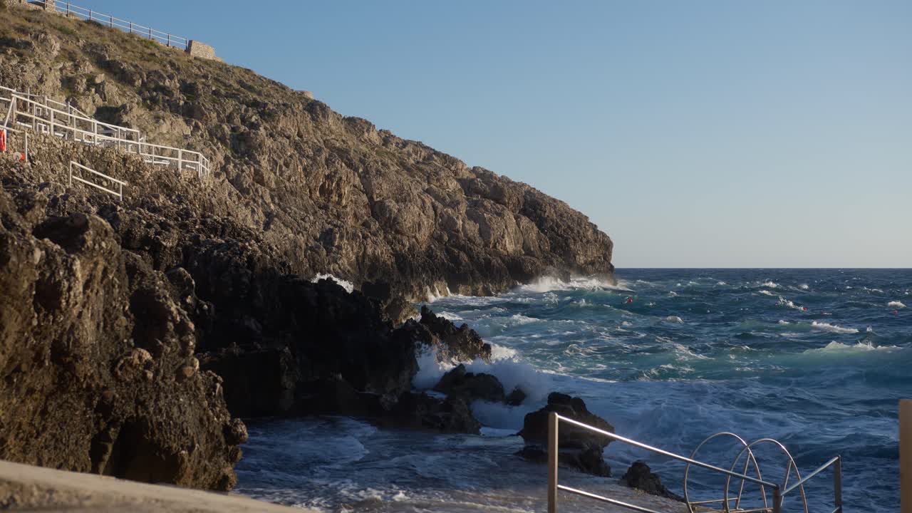 Wide shot of ocean waves crashing against rocks during sunset, with warm golden light at Faro di Punta Carena, Capri