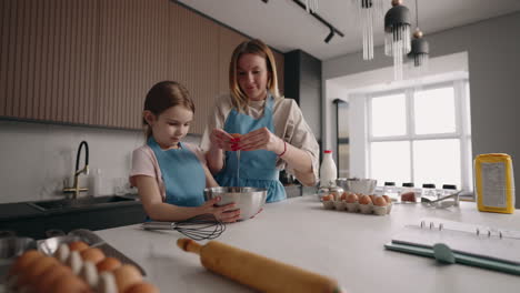cheerful mother and daughter are cooking dough or omelette in home kitchen in morning beating eggs into bowl