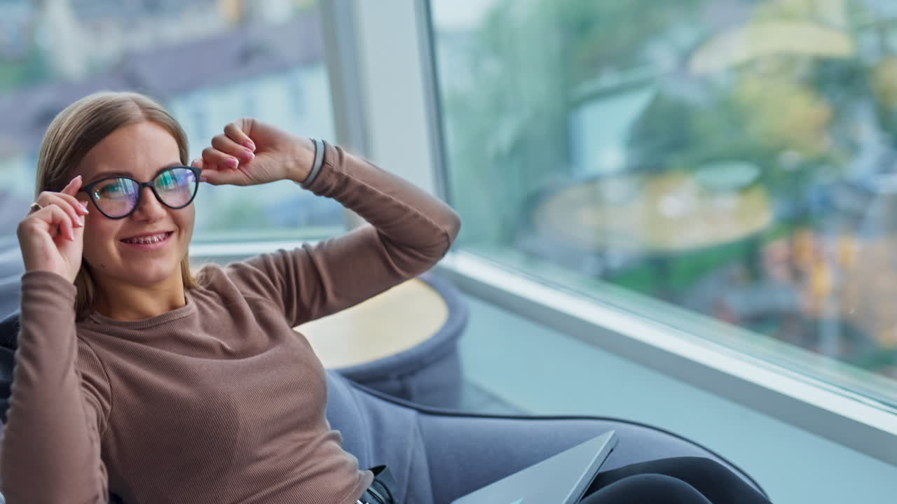 Happy smiling lady touching her glasses. Relaxed woman sitting in bean bag chair resting from work. Blurred backdrop.