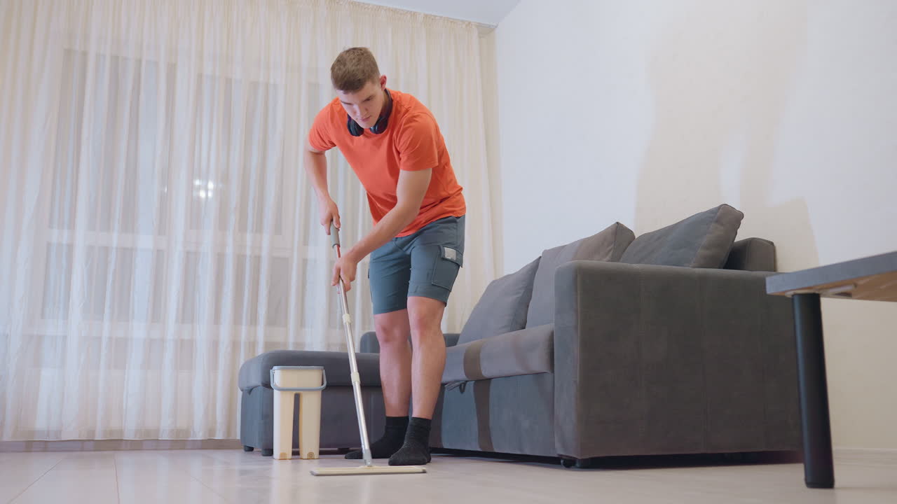 Young boy in orange shirt and shorts with headphones around neck bends forward holding mop handle while cleaning floor near gray sofa in bright living room with curtains