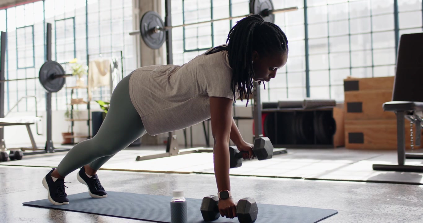 Mid-adult African woman in leggings doing plank rows with dumbbells in gym, kneeling, sipping water