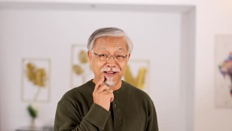 Elderly Asian man stands in a bright, modern room, pausing thoughtfully with his hand on his chin. Soft, even lighting and static camera