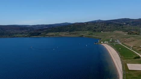 Gente-Disfrutando-De-La-Playa-Del-Lago-Con-Jardines,-Veleros-Navegando-En-Una-Tarde-Soleada