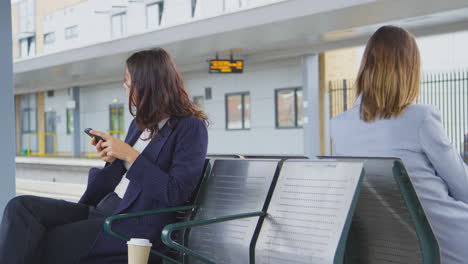 two businesswomen commuting to work waiting for train on station platform looking at mobile phones