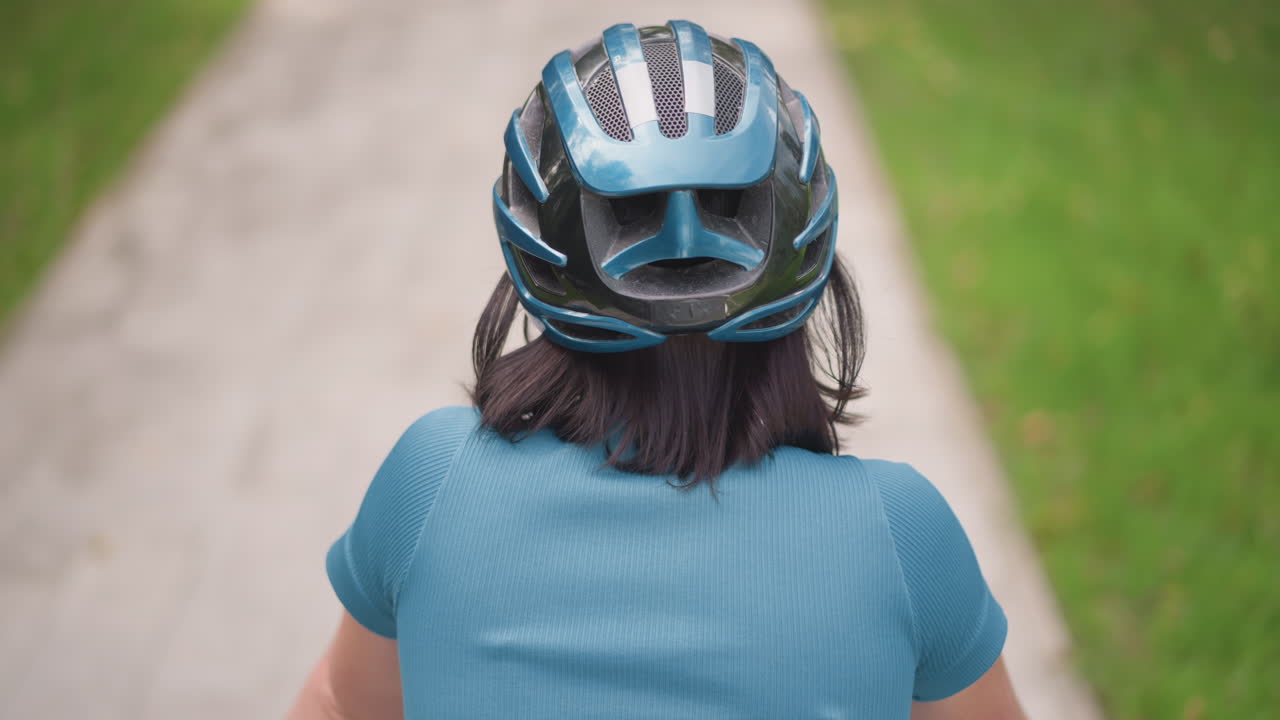 Ciclista visto da dietro su un sentiero del parco con casco blu e maglia color ottanio, capelli scuri corti e postura stabile, passerella pavimentata fiancheggiata da erba e alberi, passo lento e costante e quieta solitudine, pendolare quotidiano