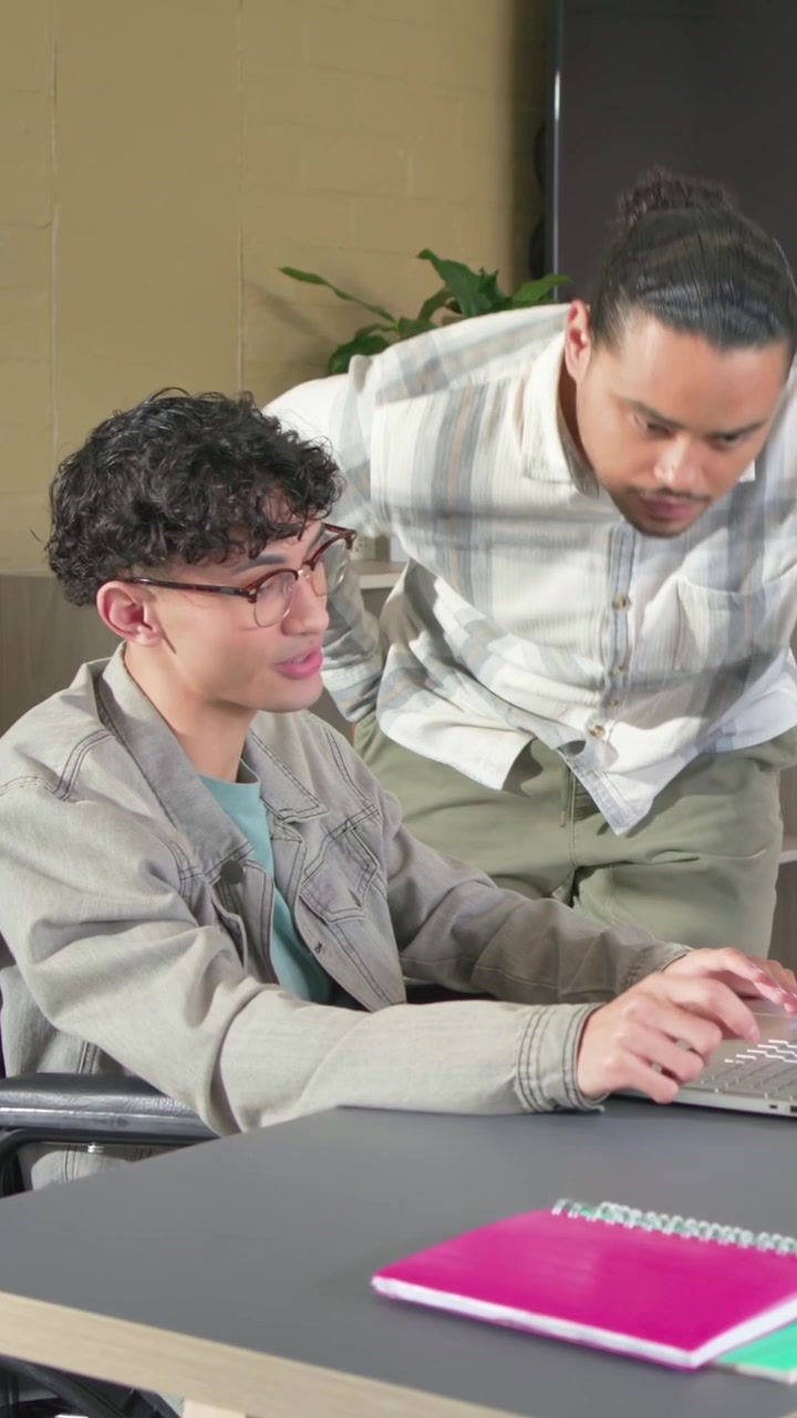 Video verticale: Entrando con un notebook, un collega si china sulla scrivania aiutando un utente in sedia a rotelle al laptop.