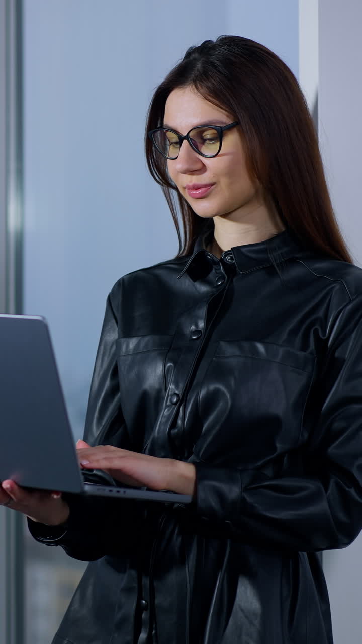 Young female business person wearing glasses types on laptop holding it in her hands. Woman finishes work, closes computer and turns to look at the window. Vertical video