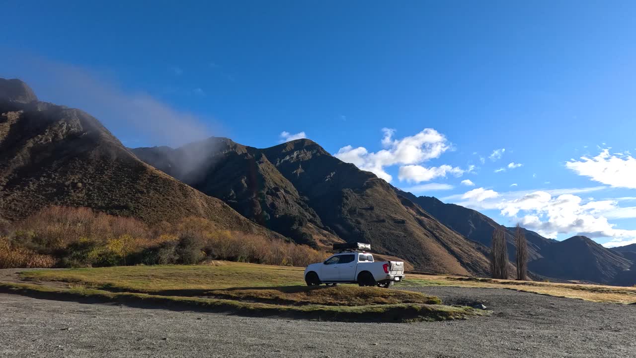 Person walks to vehicle and drives away, wide shot, bright daylight, dramatic mountain backdrop