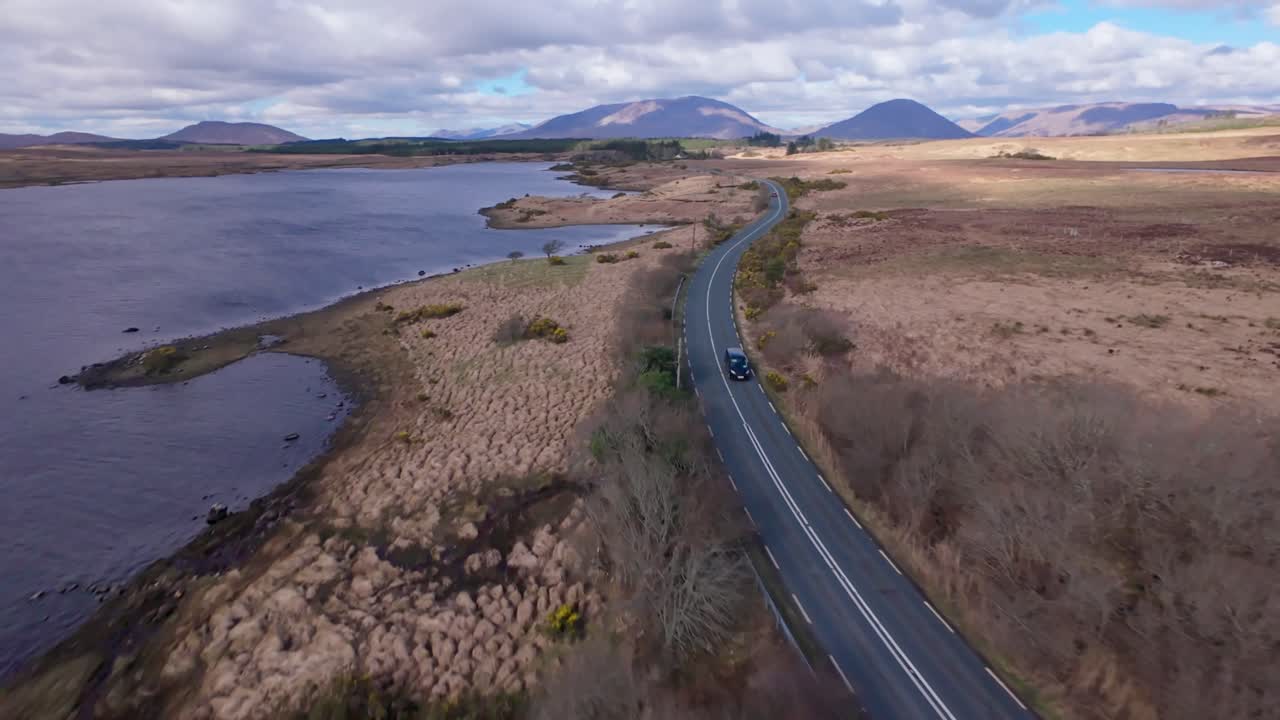 A winding road in ireland beside a serene lake with mountains in the distance, aerial view