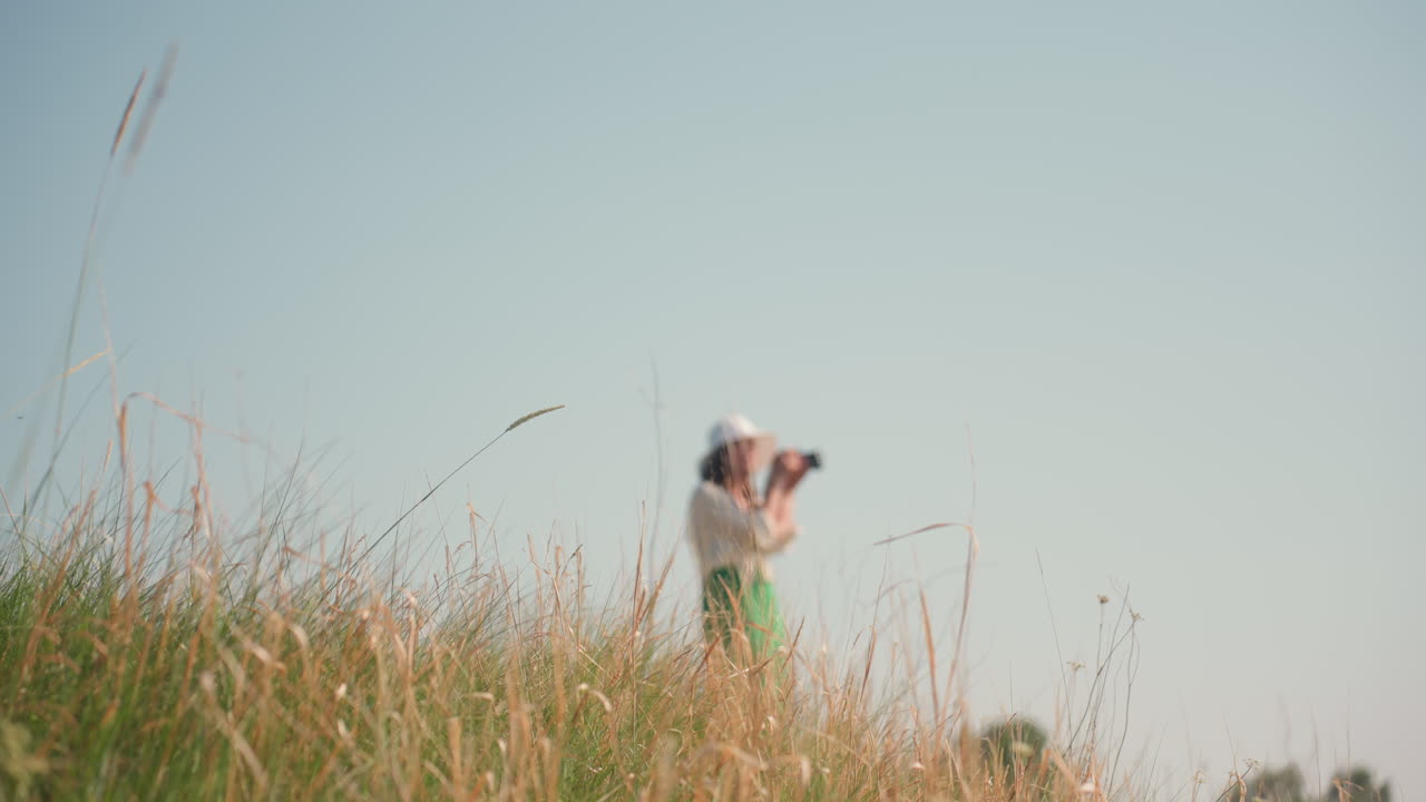 Dry golden grass in foreground moves gently in sunlight while blurred woman wearing white hat and green skirt stands in distance raising camera under blue sky