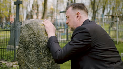 close up view of man in black suit kneeling in front of a tombstone and touching it 1