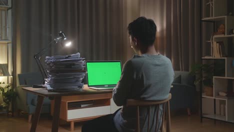 vista trasera de un hombre asiático haciendo una llamada de video con una computadora portátil de pantalla verde mientras trabaja con documentos en casa