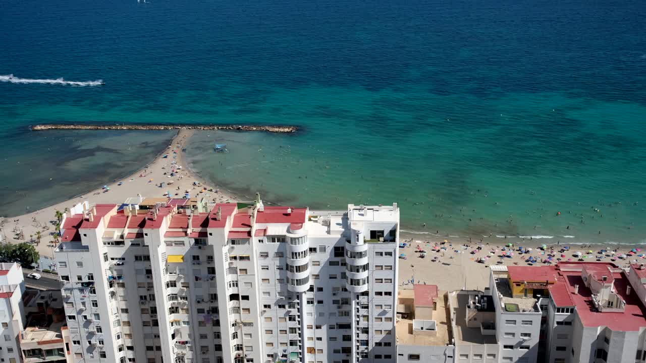 A breathtaking top view showcases towering high-rise buildings, crystal-clear blue ocean waters, and a pristine white sandy beach, blending urban and coastal beauty.