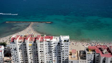a breathtaking top view showcases towering high-rise buildings, crystal-clear blue ocean waters, and a pristine white sandy beach, blending urban and coastal beauty.