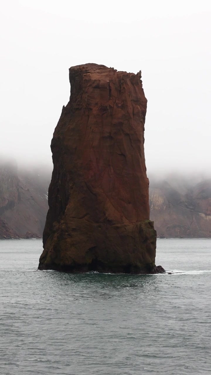 Dramatisk vertikal video fanger den tårnhøje havstabel Raven Rock, der dukker op fra tågede antarktiske farvande ved indgangen til Deception Island, med tågeindhyllede klipper og overskyet naturligt lys.