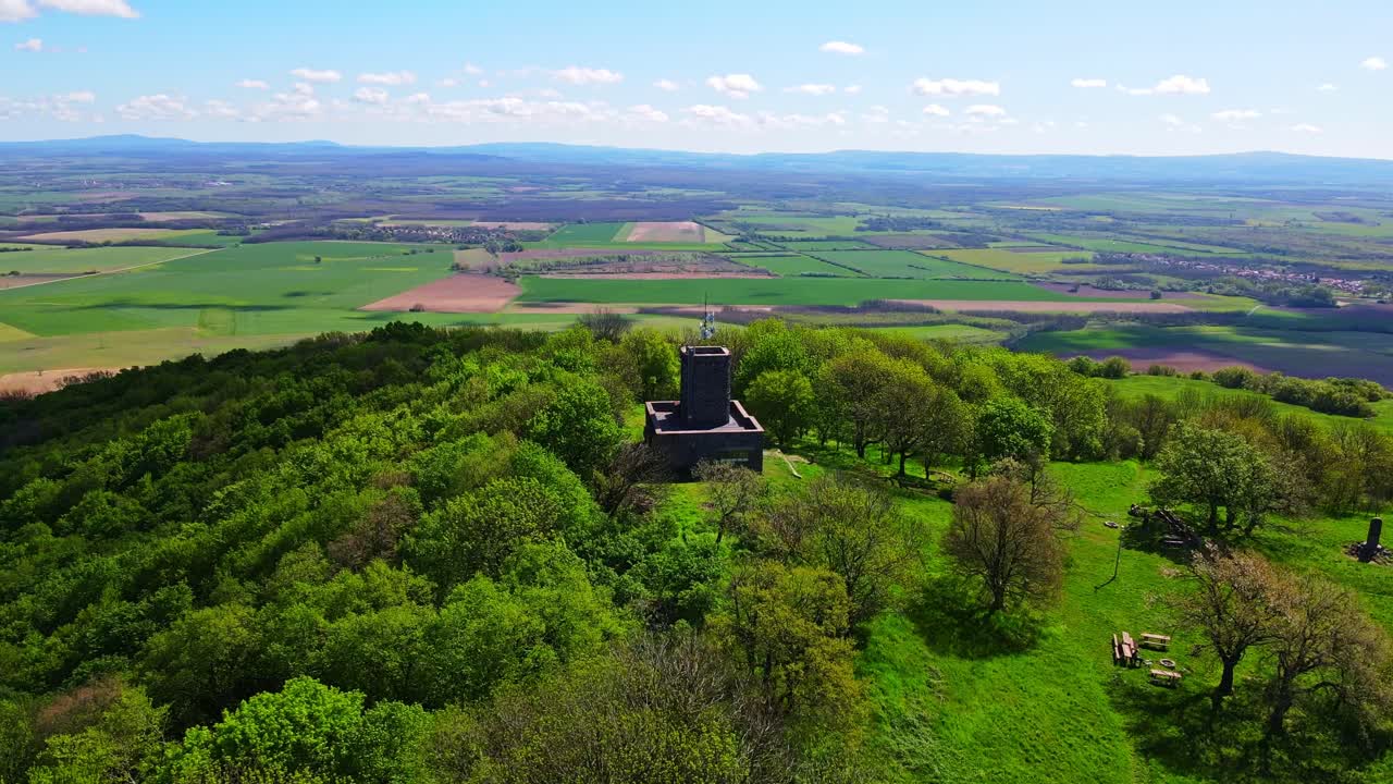 An aerial drone video shows a historic lookout tower surrounded by lush green trees atop a small hill, with vast agricultural fields and distant mountains stretching into the horizon under a blue sky