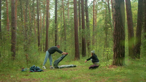 young man folds tent fabric on forest ground while woman in beanie squats nearby watching, surrounded by tall trees, soft grass, and scattered camping gear during peaceful outdoor adventure