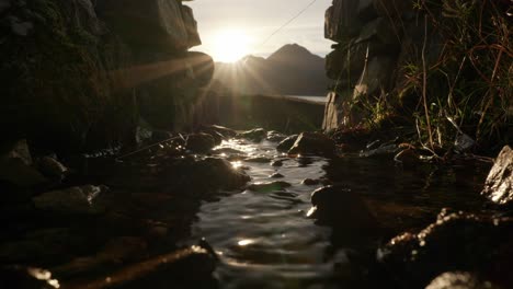 the the sun sets behind a mountain to produce a sunburst in the background as a small river gently flows under an arch in a stone wall