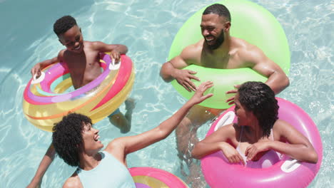 Felices-Padres-Afroamericanos,-Hijo-E-Hija-Flotando-En-Inflables-En-Una-Piscina-Soleada,-Cámara-Lenta