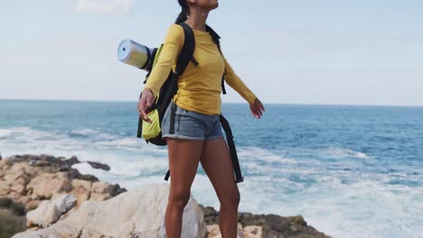 african american woman with backpack standing with her arms wide open near the ocean while hiking