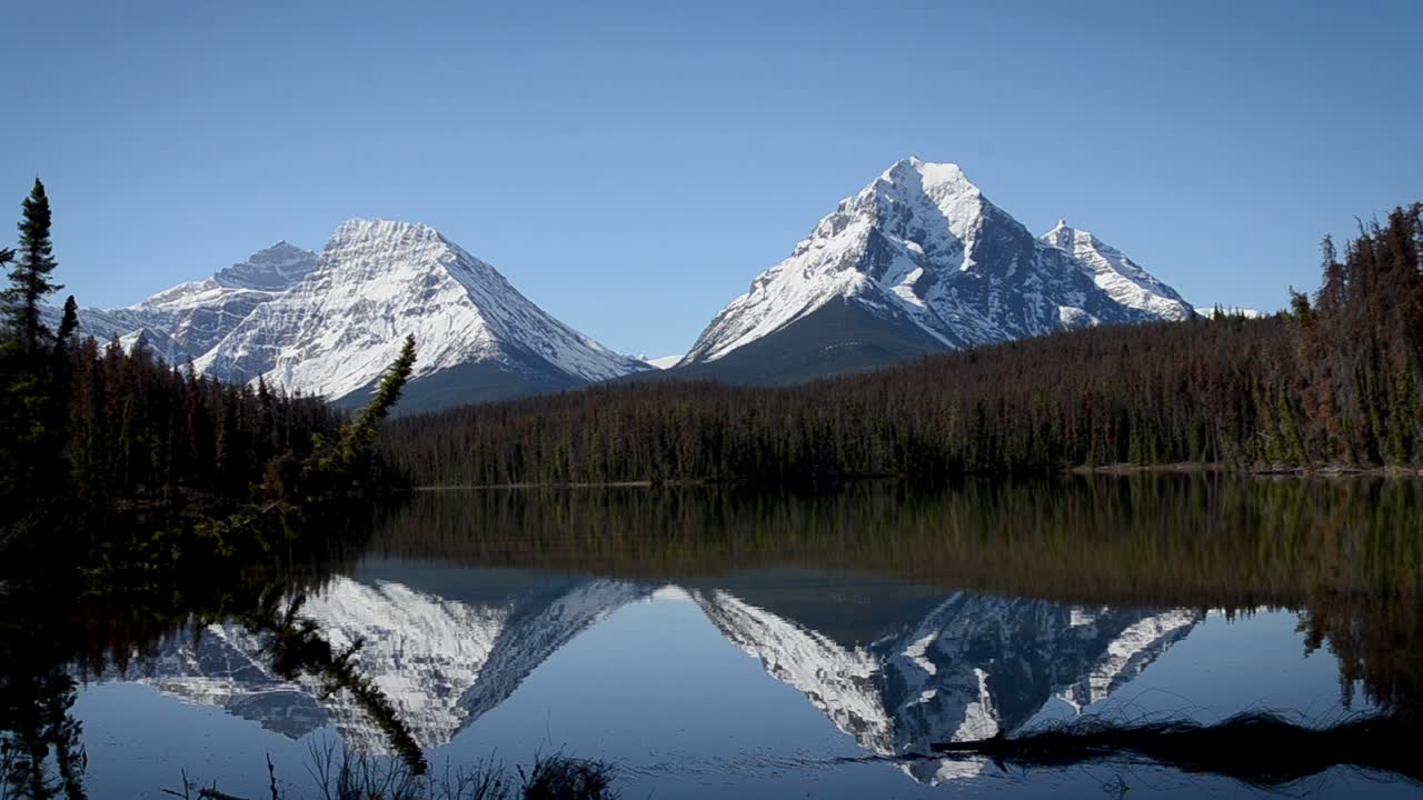 Three Sisters Peaks reflect in calm surface of Policemans Creek near Canmore as clear sky and snowy summits create crisp mountain scene captured through steady timelapse tripod perspective