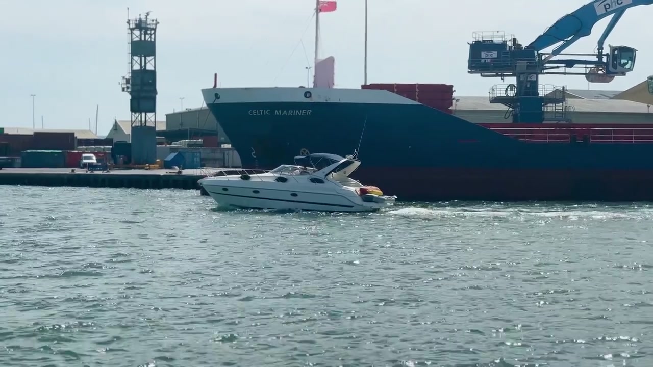 Cargo ship and leisure boats moving across Poole Harbour with working port cranes and shimmering water under bright daytime sky in Dorset, England, United Kingdom