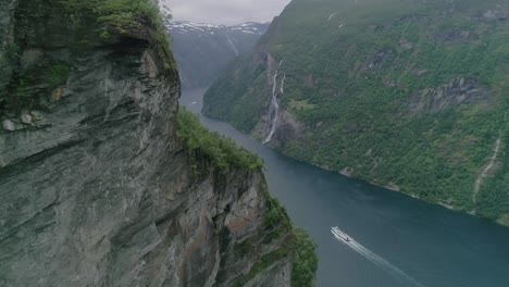 Slomo-Luftaufnahme-Beim-Flug-In-Richtung-Des-Geiranger-Fjords-Mit-Einem-Fahrenden-Boot-Und-Dem-Wasserfall-Der-Sieben-Schwestern-Im-Hintergrund