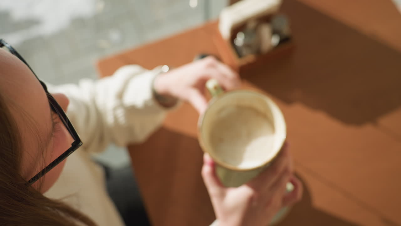 Close up of student holding ceramic coffee cup above saucer with spoon on wooden table, earbud case and condiments in background