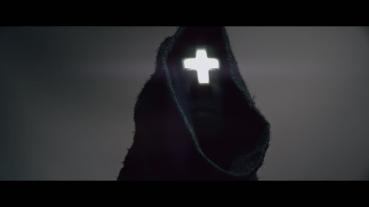 Masked and hooded man with cross of light glowing on his face looks in the camera