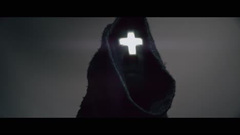 masked and hooded man with cross of light glowing on his face looks in the camera
