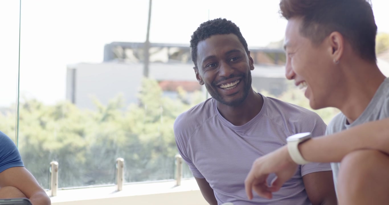 Diverse male friends sitting on mats near window, left man showing tablet, passing bottle, laughing