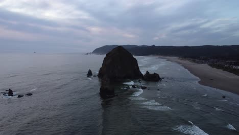 haystack rock silhouette at sunset in cannon beach, oregon from high above. zoom shot filmed with 4k drone as the waves roll onto the beach and the mountains peak out in the background.